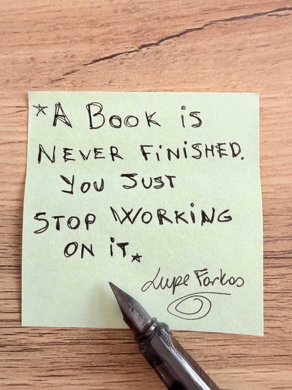 Handwritten note saying “A book is never finished, you just stop working on it” with a pen on a wooden table